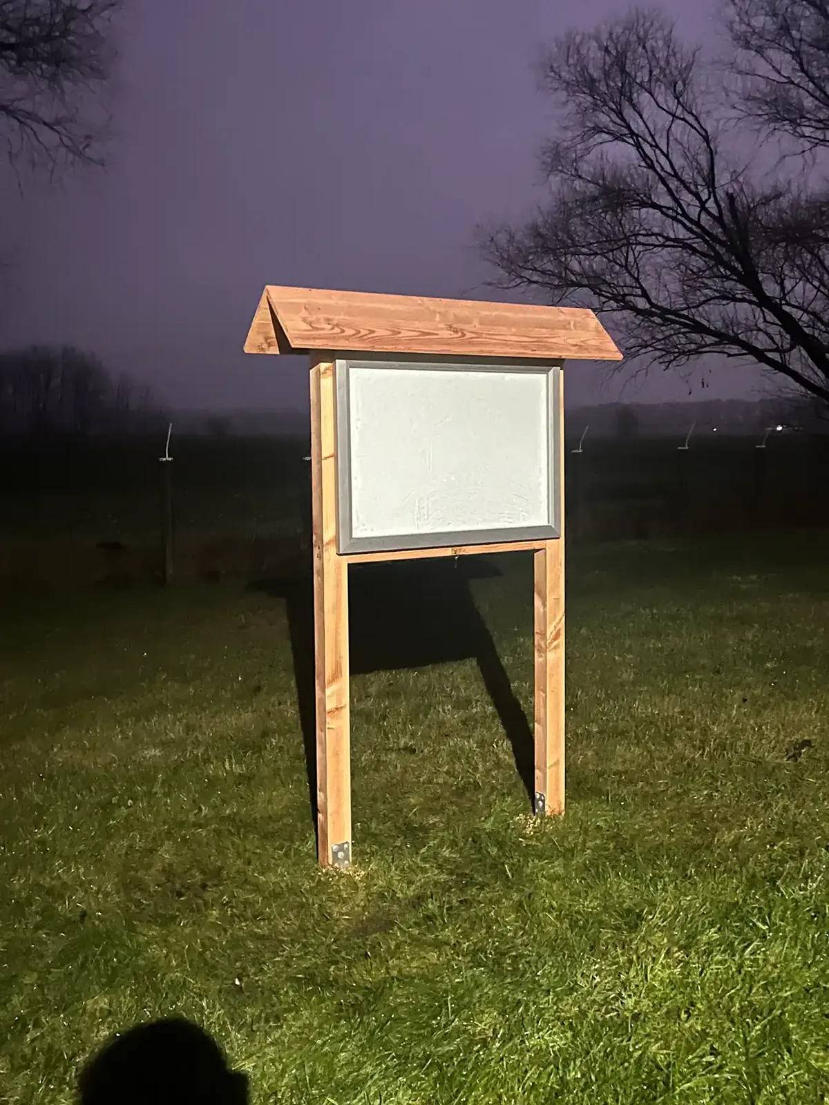 Freistehender Schaukasten im Holzgestell mit Dach und heller Aushangfläche auf einer Wiese bei Dämmerung.